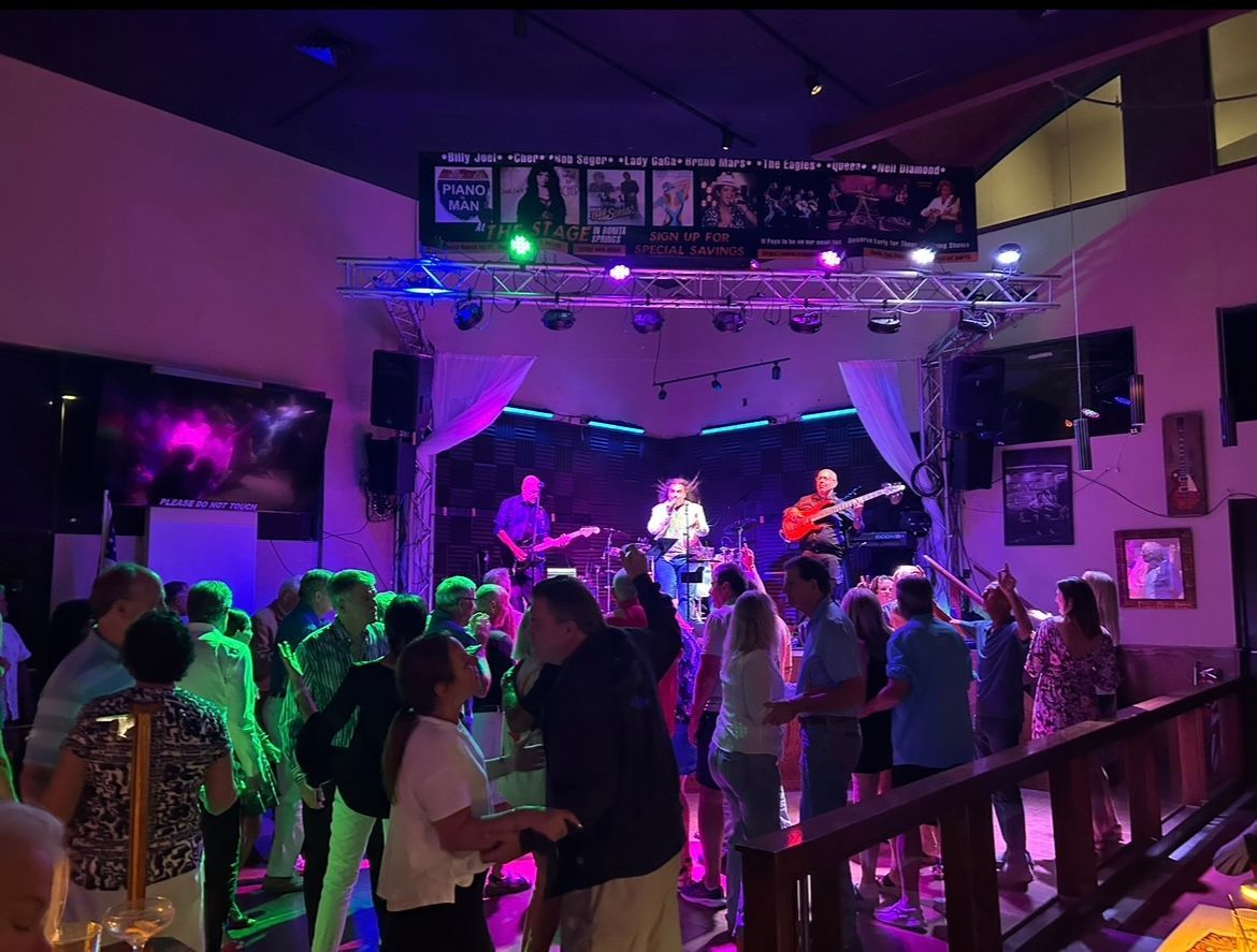 Crowded nightclub with live band on a lit stage and people dancing in purple-green light