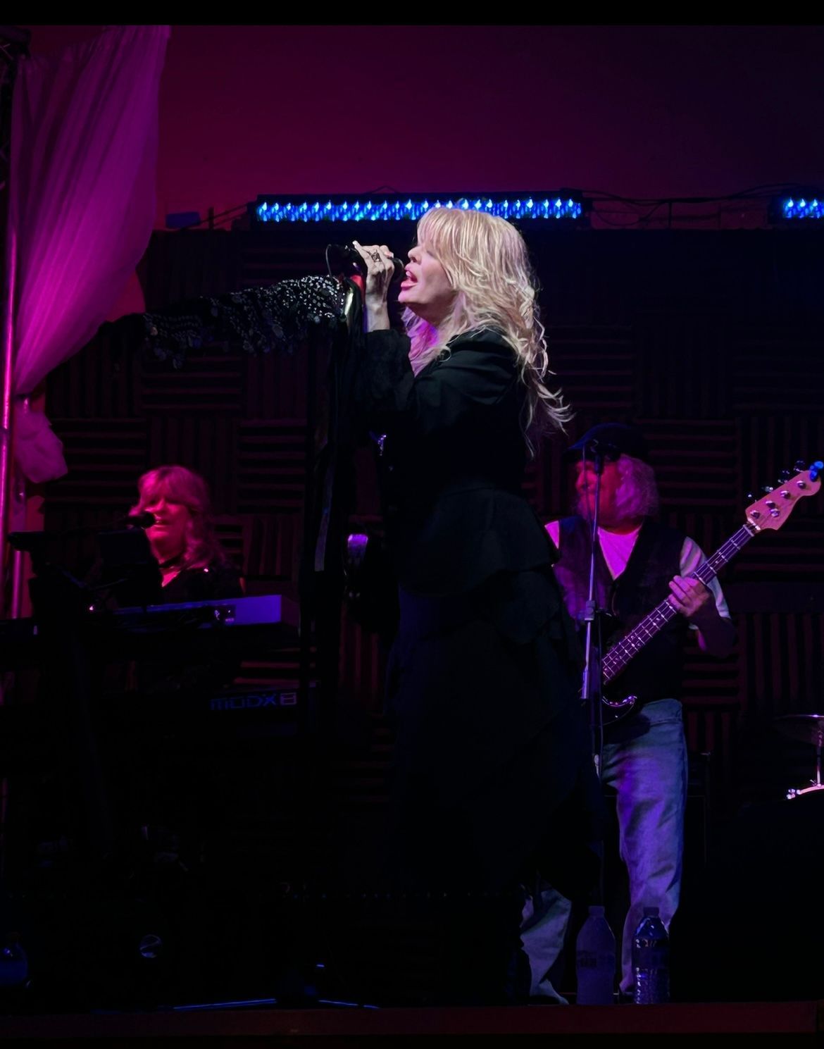 Blonde singer in black coat performing onstage with keyboardist and bassist under purple lighting