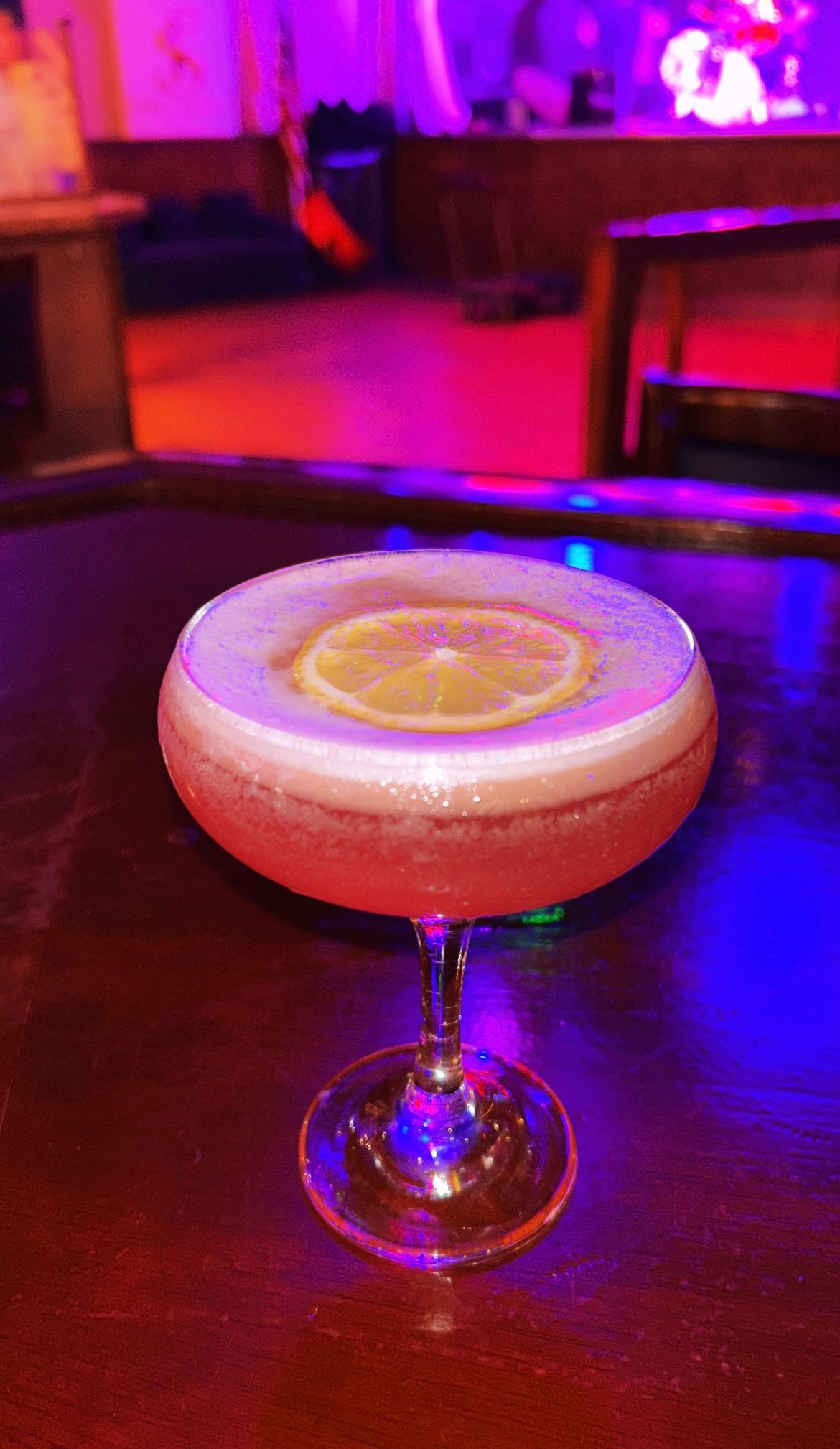 Pink cocktail in a coupe glass with a lemon slice, set on a table in a dimly lit bar.