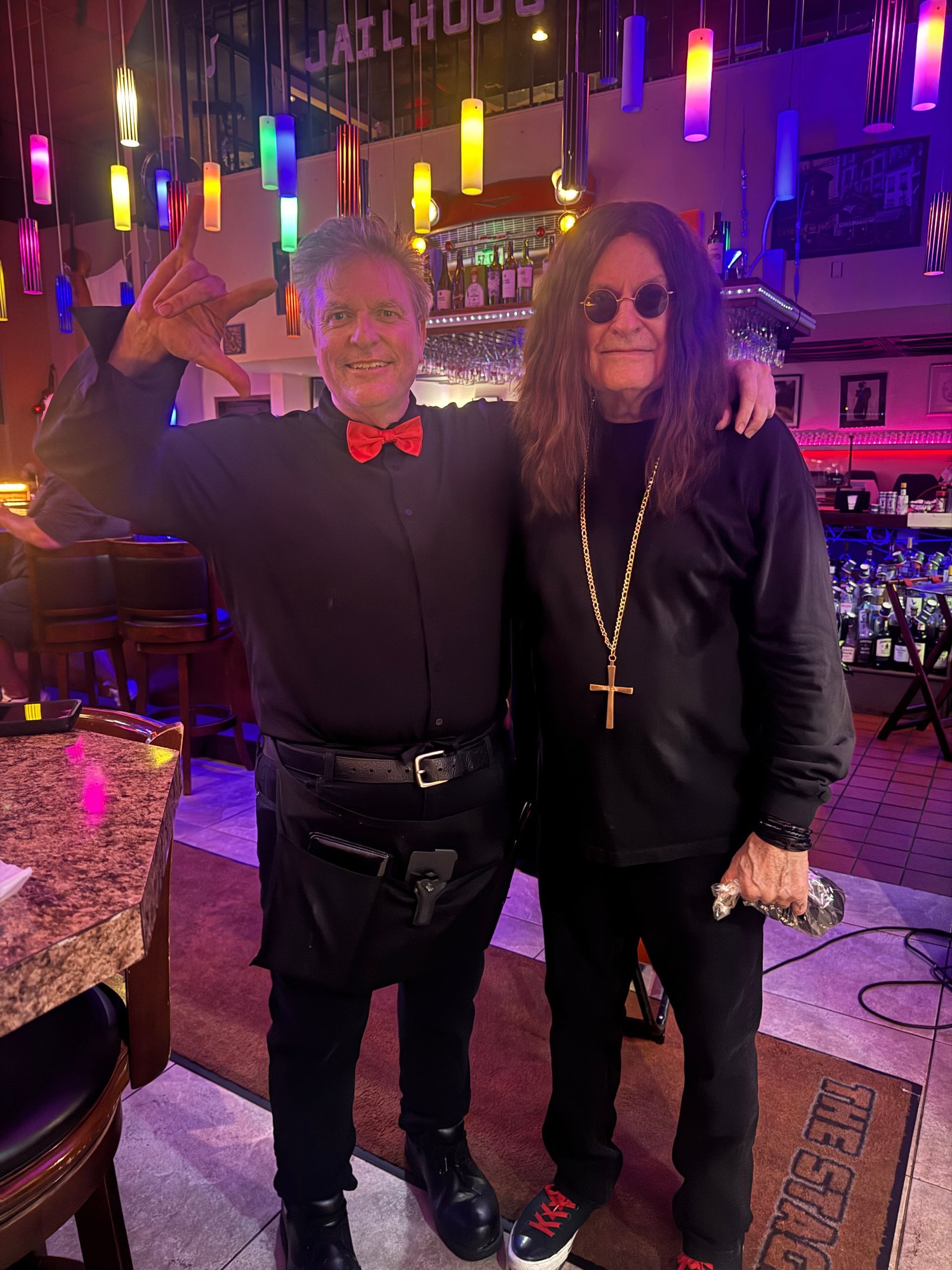 Two people posing in a colorful nightclub, one waving and one holding a cross, under neon lights.