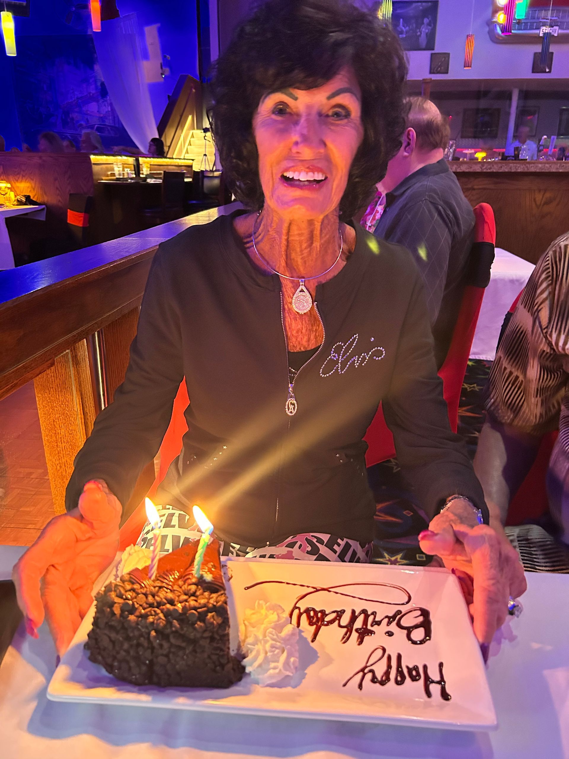 Woman smiling at a table with a birthday cake and lit candle in a dimly lit restaurant