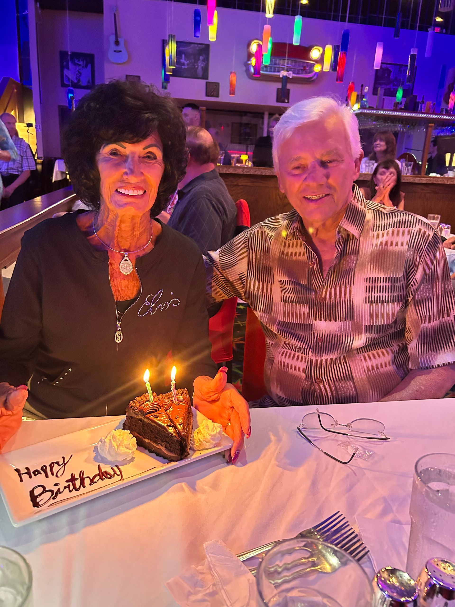 Two people smiling at a restaurant table with a birthday cake and lit candle.