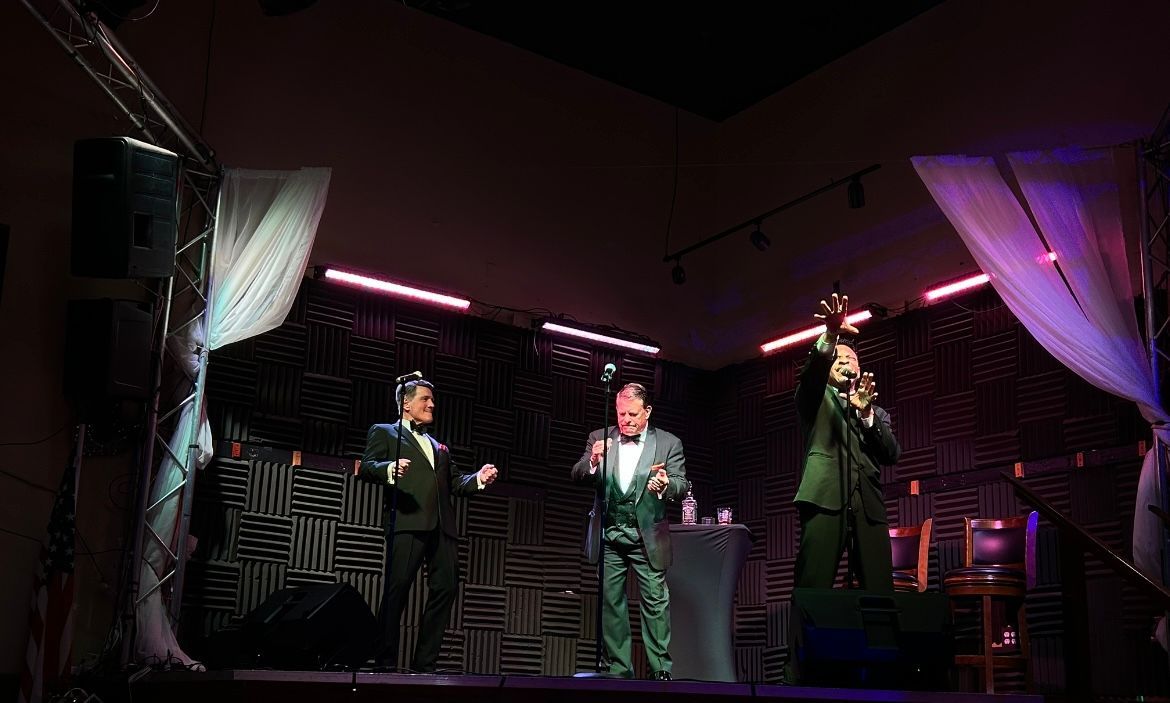 Three performers on a stage under pink and purple lights, each holding a microphone.