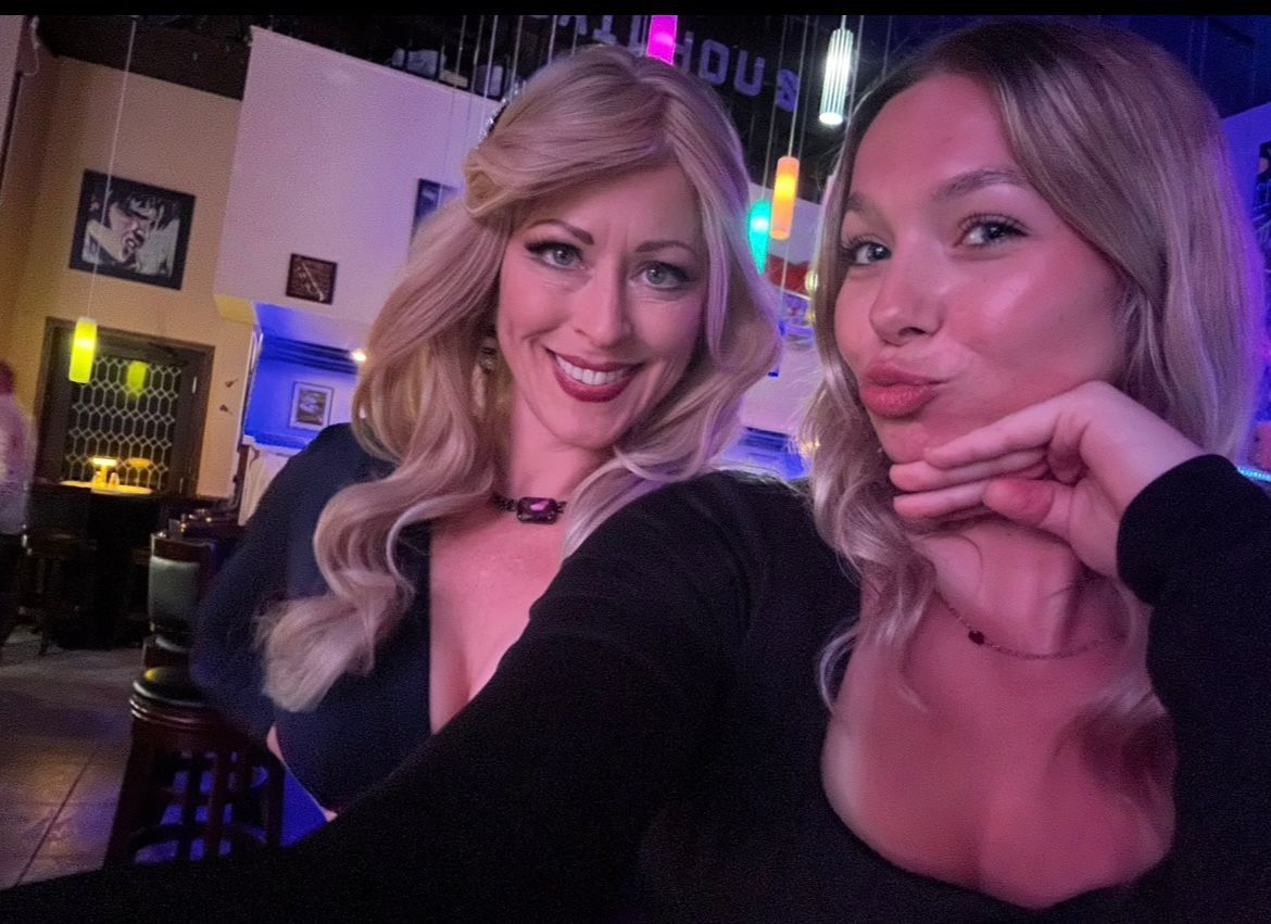 Two people smiling and posing for a selfie in a dimly lit bar with colorful neon lights