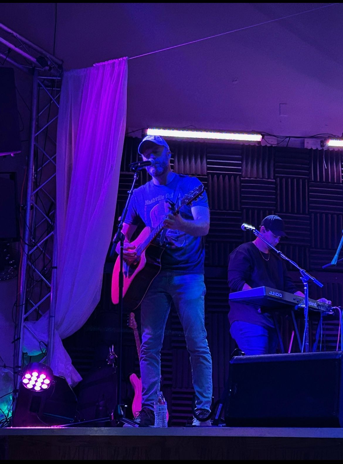 Musicians performing on a purple-lit stage, one playing guitar and one seated at a keyboard.