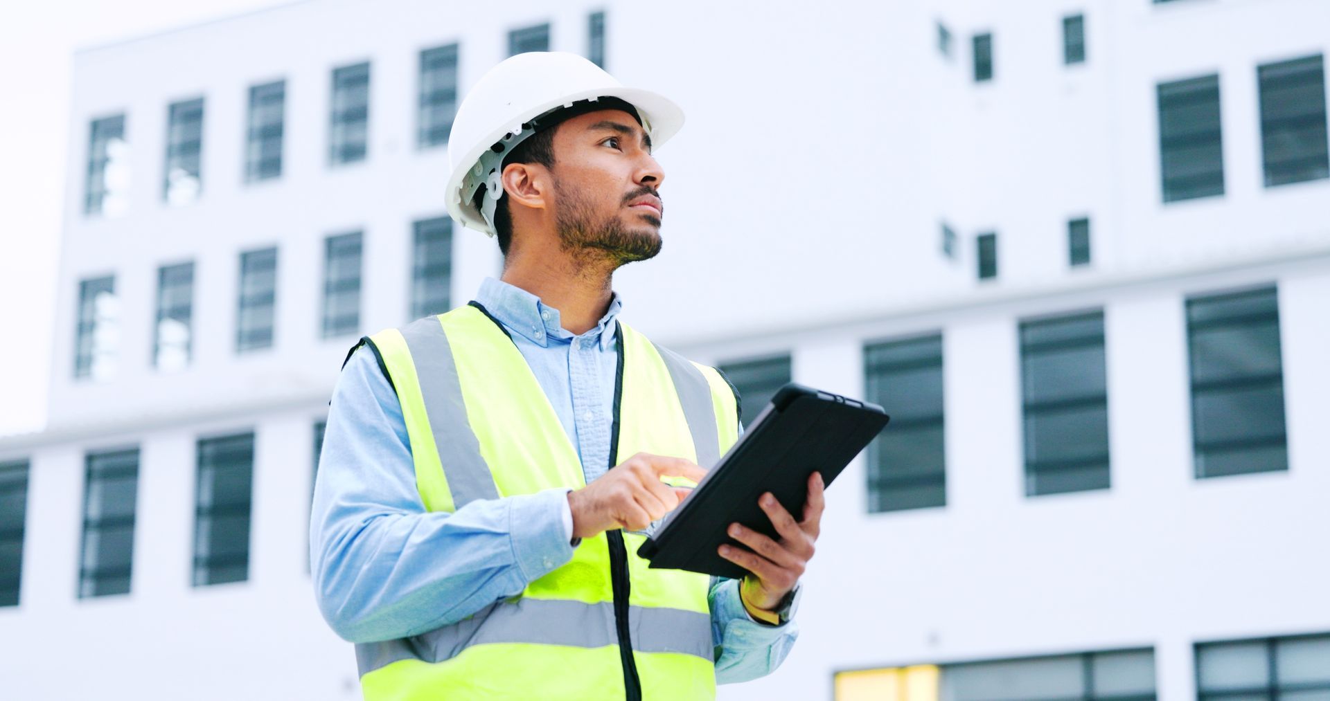 A person in a hard hat and high-visibility vest holds a tablet while standing in front of a modern white building.