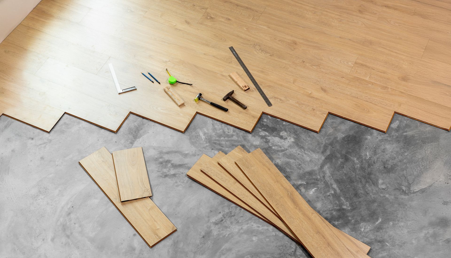 Light-colored laminate floor planks partially installed over a concrete subfloor, with hand tools arranged on top.
