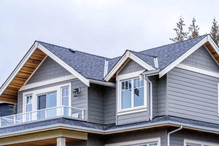The upper exterior of a gray, two-story house featuring gabled roofs, wooden soffits, and a small balcony.