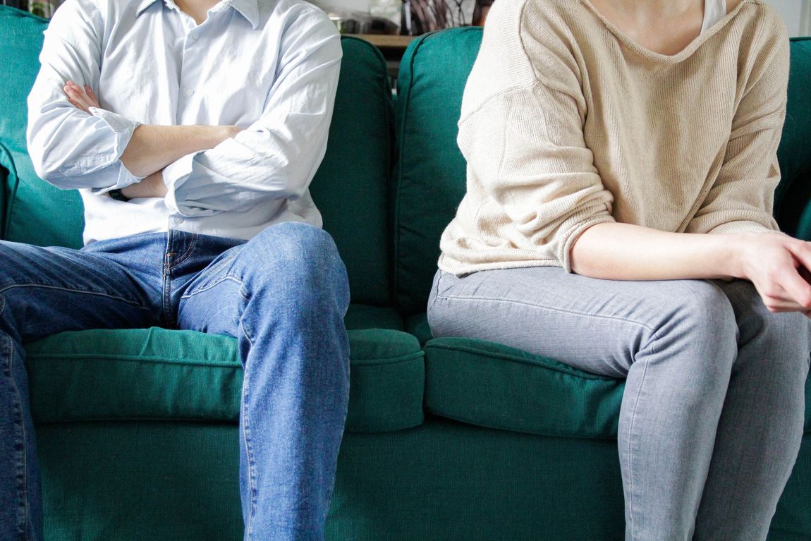 Un uomo con le braccia incrociate e una donna seduti separatamente su un divano verde, che guardano altrove.