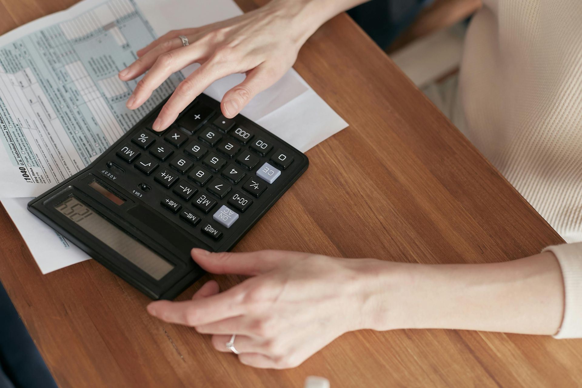Person's hands using a calculator on a wooden table, with paperwork visible.