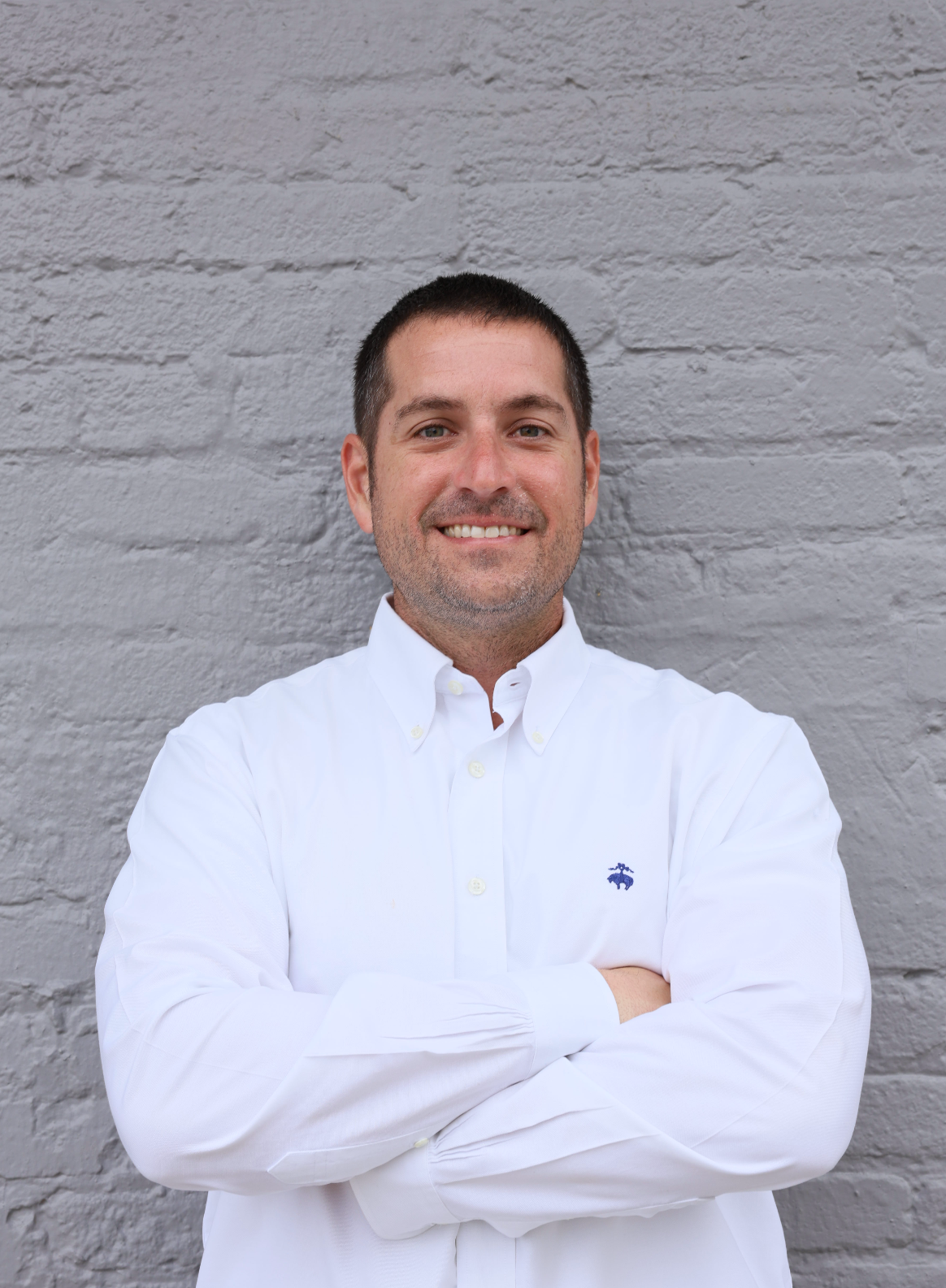 A man in a white shirt is standing in front of a brick wall with his arms crossed.