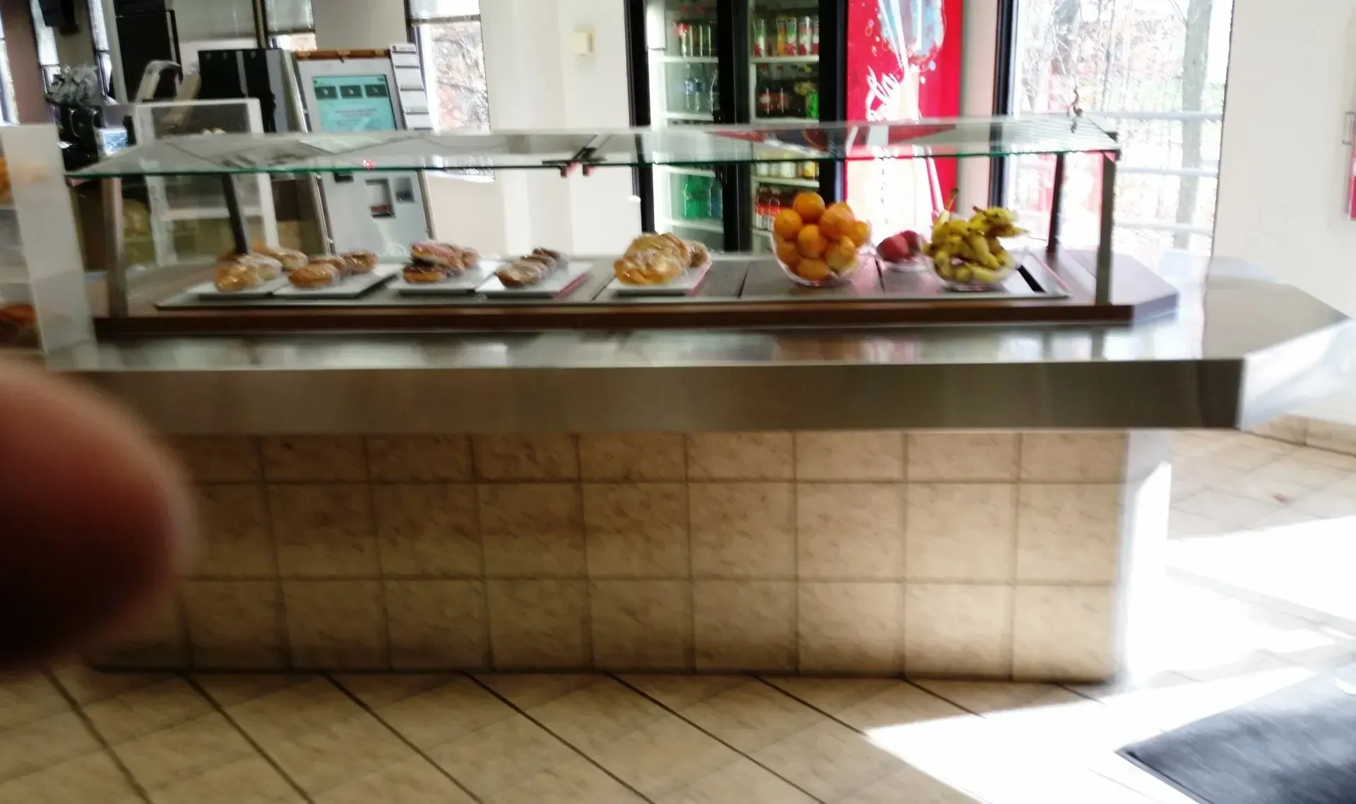 Cafeteria counter with trays of pastries and fruit behind glass, tiled front, and bright entrance light.