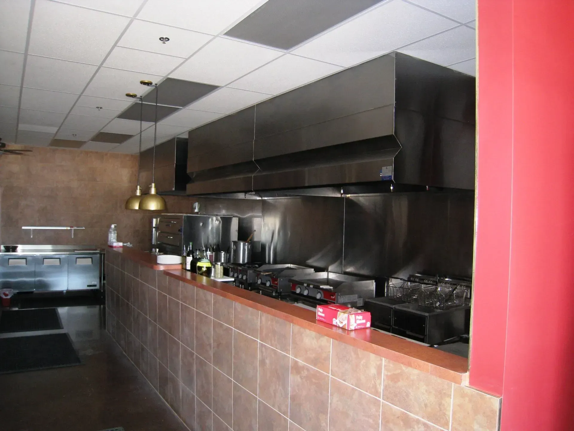 Empty restaurant kitchen with stainless steel cooking line, tiled counter, and red wall panel.