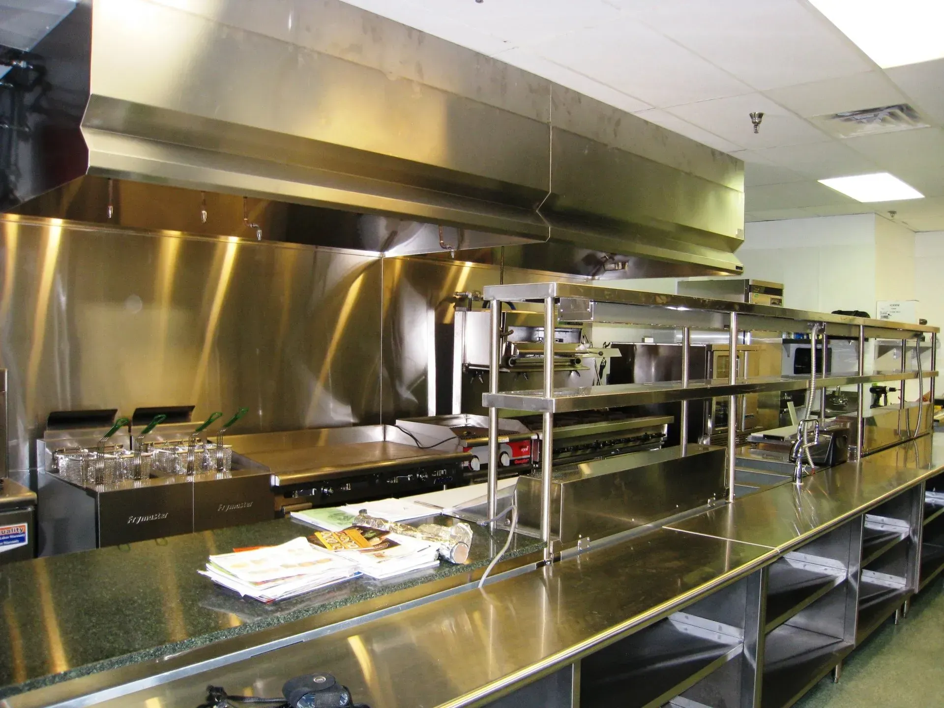 Empty stainless-steel commercial kitchen with metal counters, shelves, and a large ventilation hood