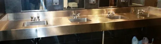 Stainless steel commercial kitchen sink and counter with multiple faucets and dishes drying underneath.