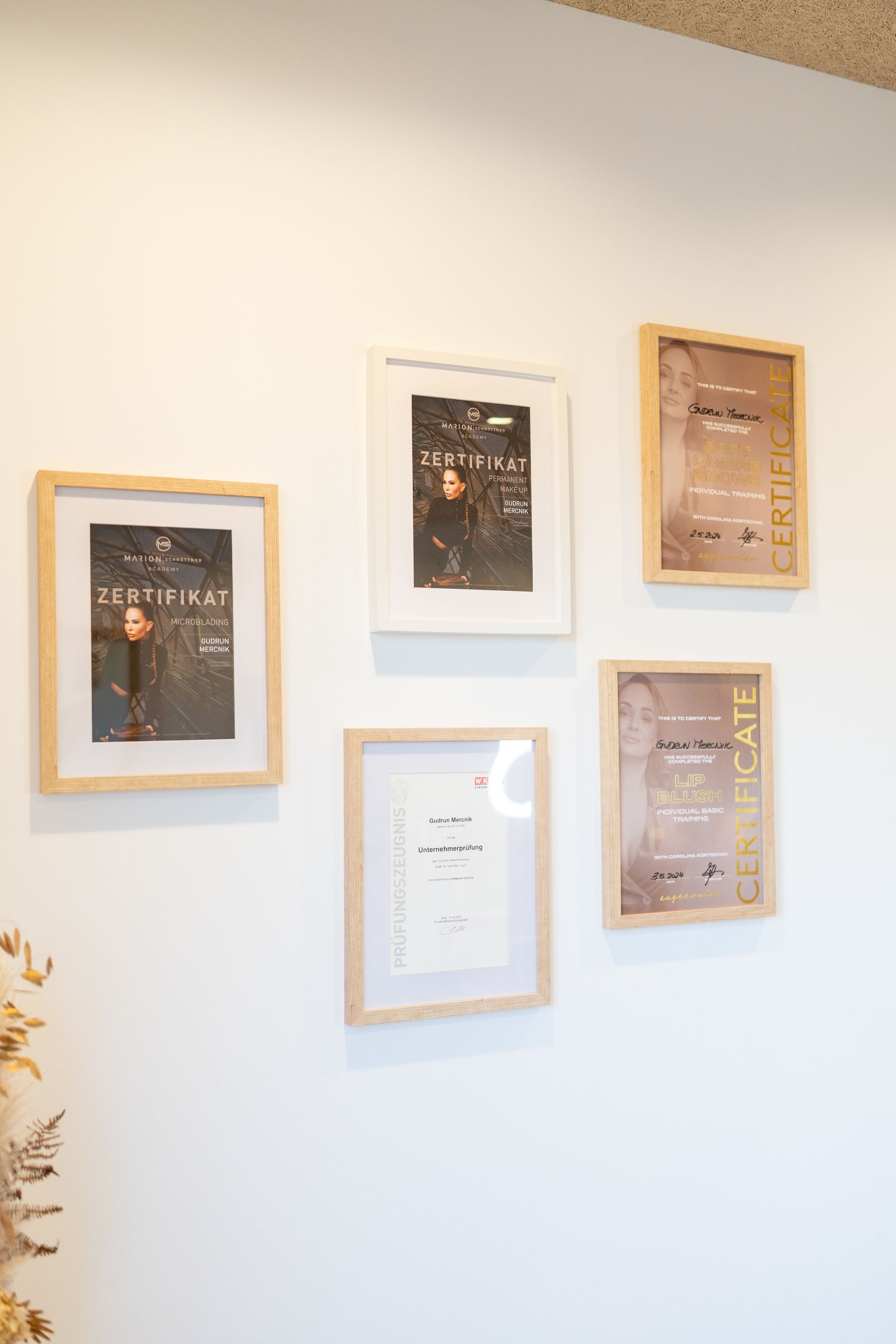 Fünf gerahmte Bilder an einer weißen Wand. Die Rahmen sind aus hellem Holz oder weiß und enthalten Bilder und Urkunden.