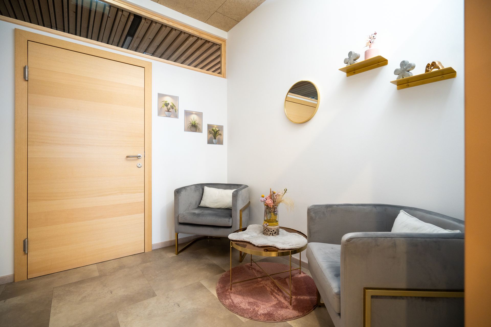 Gemütlicher Wartebereich mit grauen Sesseln, rundem goldenen Tisch, rosa Teppich, Holztür und Kunstwerken an weißen Wänden.