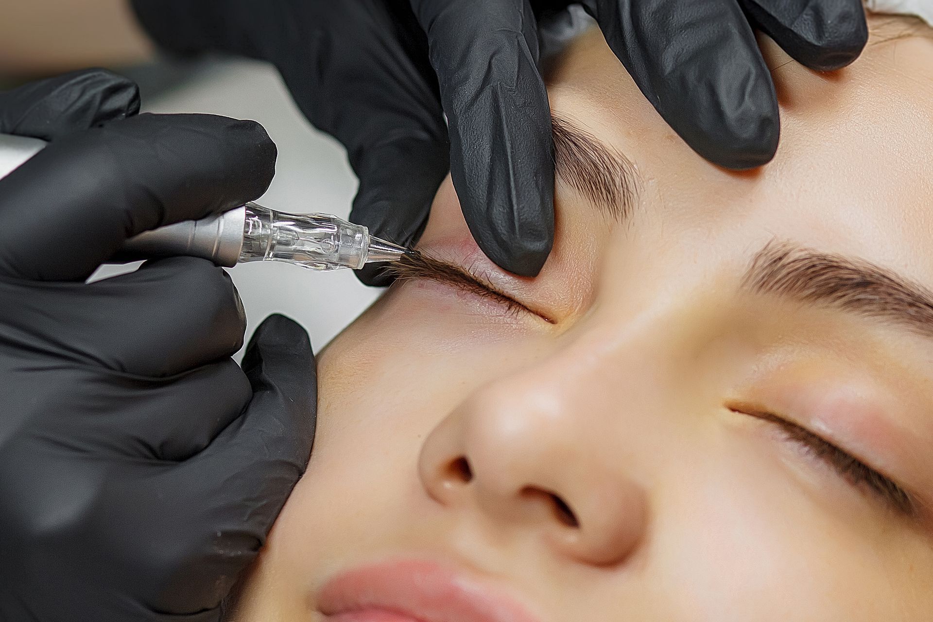 Person, die sich mit einem von behandschuhten Händen gehaltenen Gerät ein Eyeliner-Tattoo stechen lässt.