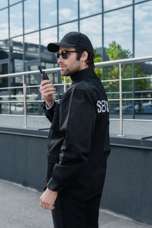 Man in black jacket and cap holding a walkie-talkie on a rooftop terrace