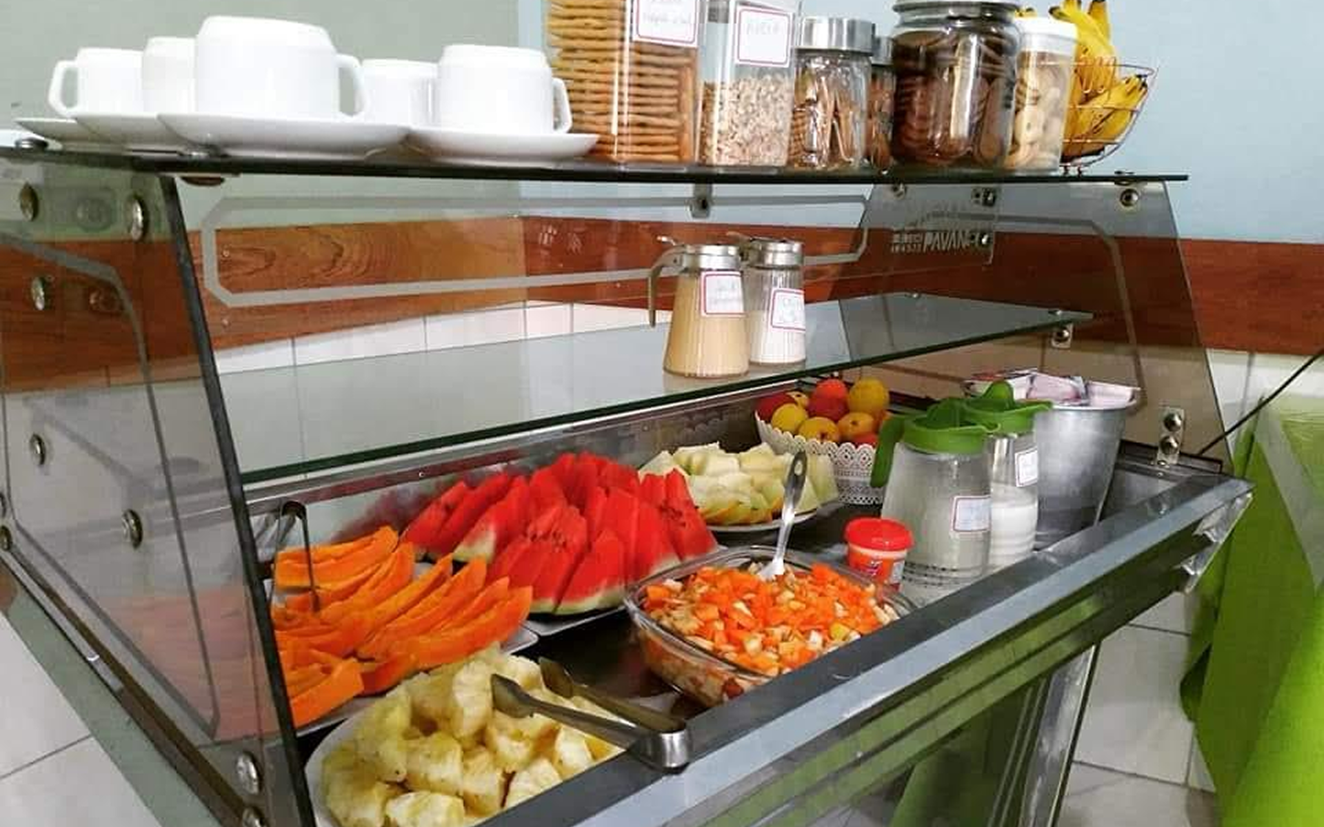 Buffet de café da manhã com frutas, doces e condimentos em vitrine refrigerada.