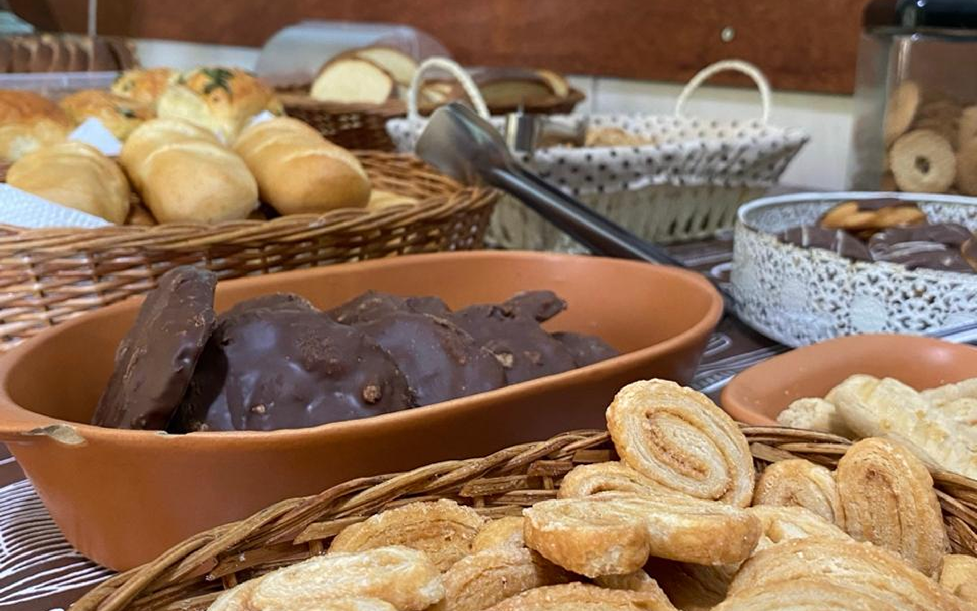 Diversos produtos de panificação em cestas e tigelas sobre uma mesa, incluindo pão, biscoitos e doces.