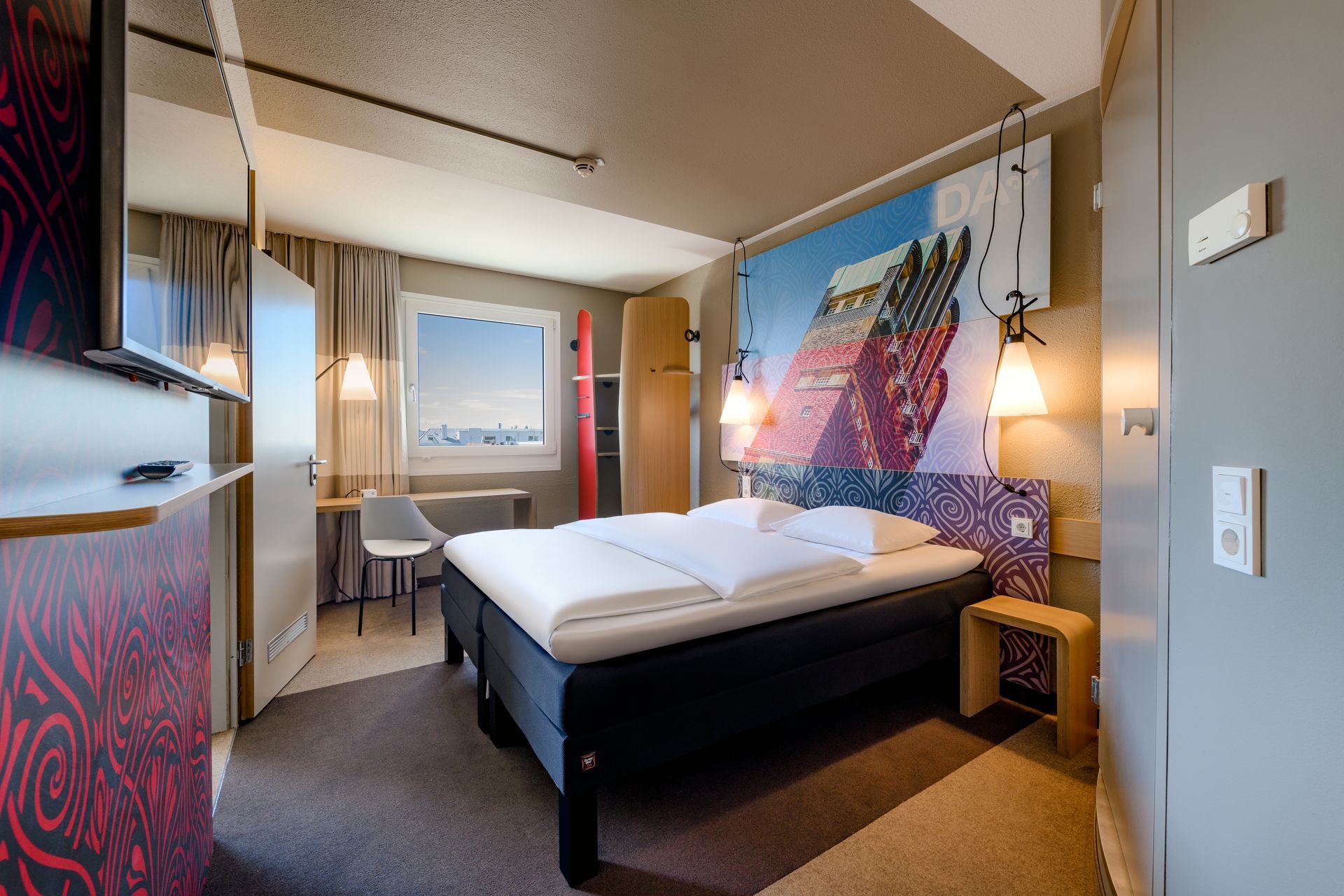 Modern hotel room with a double bed, desk by the window, and colorful wall art