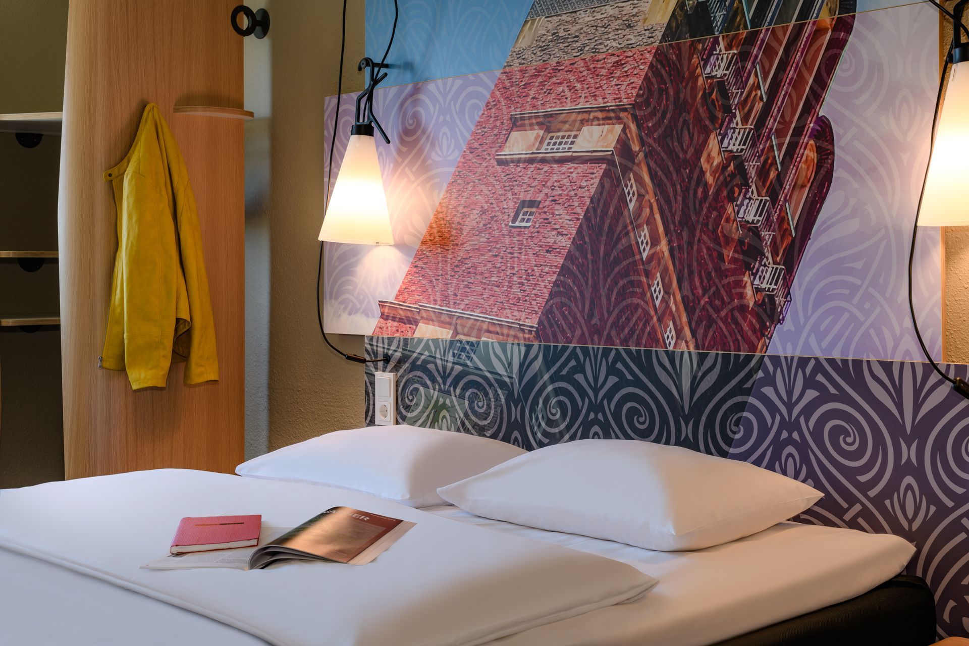Modernes Hotelzimmer mit weißem Bett, farbenfroher Wandkunst, Hängelampe und gelbem Bademantel an der Wand