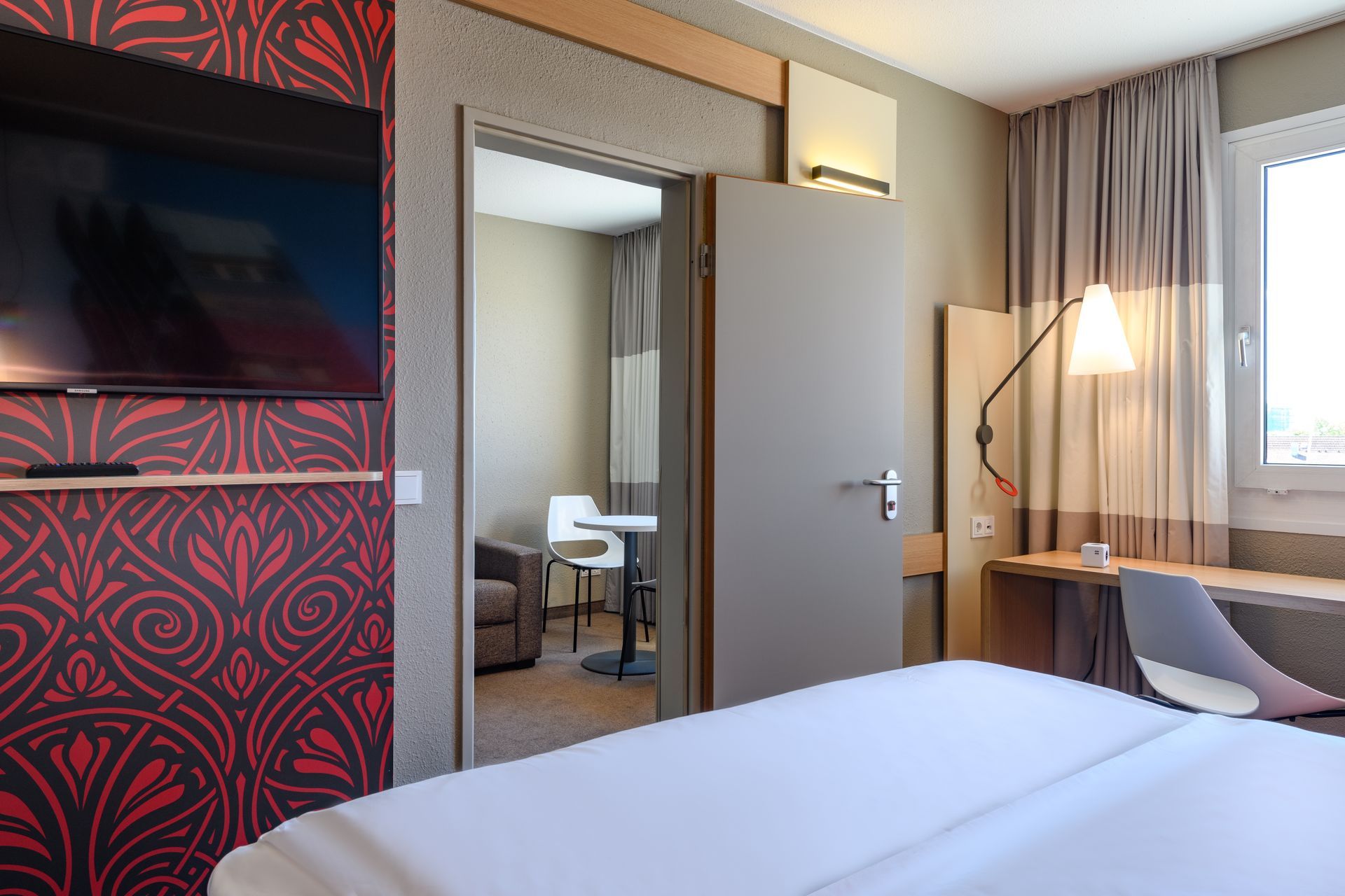 Modernes Hotelzimmer mit Bett, Fernseher, Schreibtisch, Spiegel und Fenster mit Vorhängen.