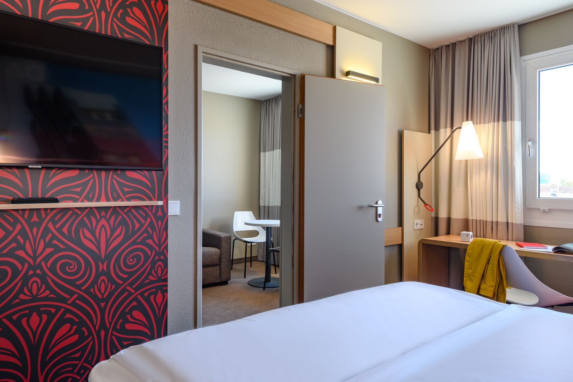 Modern hotel room with bed, wall-mounted TV, mirror, desk, and window with curtains