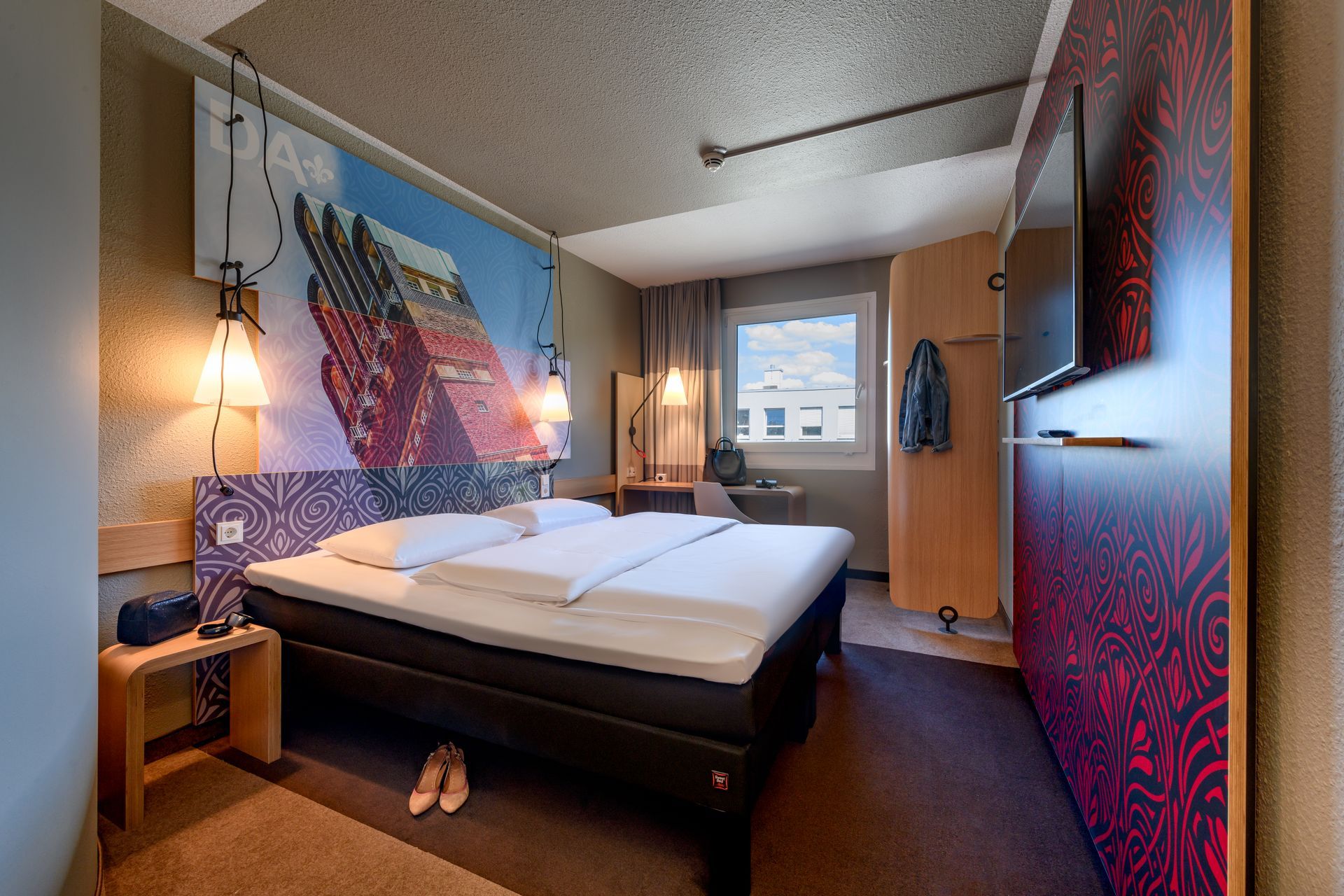 Gemütliches Hotelzimmer mit gemachtem Bett, farbenfroher Wandkunst, Nachttischlampen und Fensterblick.