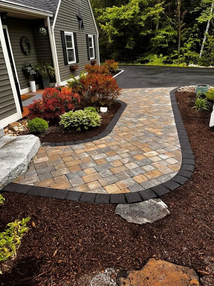 Brick walkway with dark border leads to a gray house with colorful landscaping and an asphalt driveway.