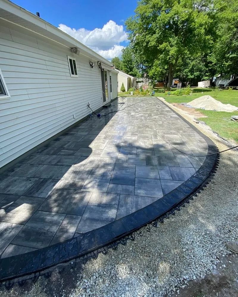 A new patio with dark gray pavers next to a white house and lawn, under a sunny sky.