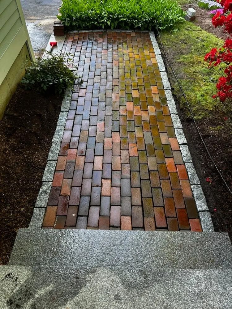 Brick pathway with steps, bordered by stone and mulch, leading to greenery.