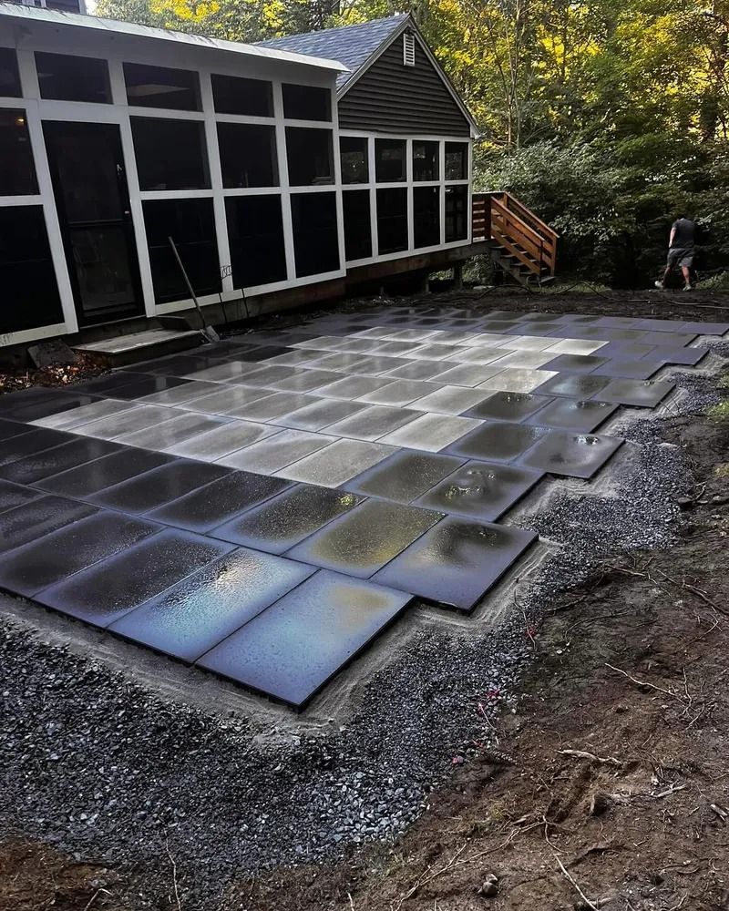 Installing dark gray square pavers on a gravel base near a cabin with screened porch; daylight.