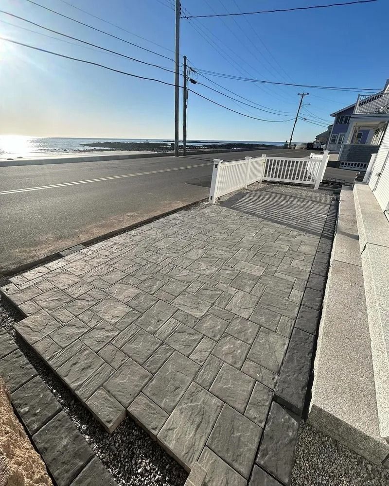 Paver walkway with white fence beside road, ocean in background. Sunny day.
