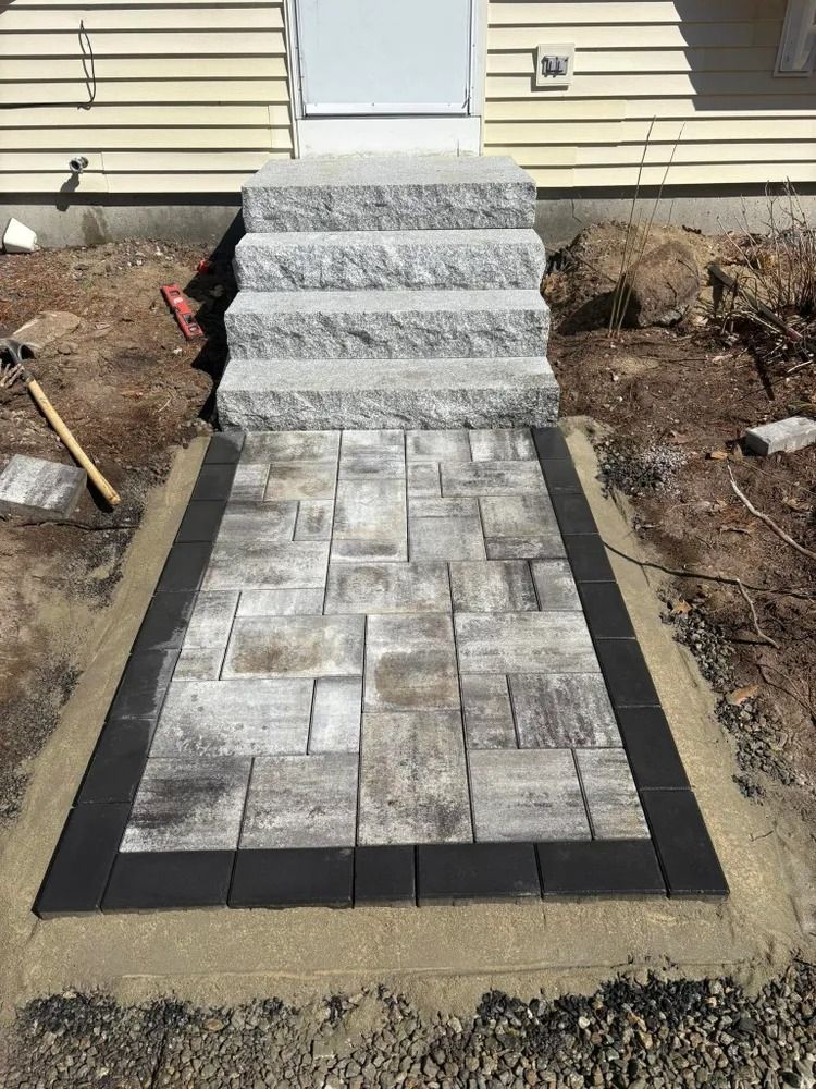 Stone steps and walkway leading to a doorway, bordered by dark pavers on concrete.