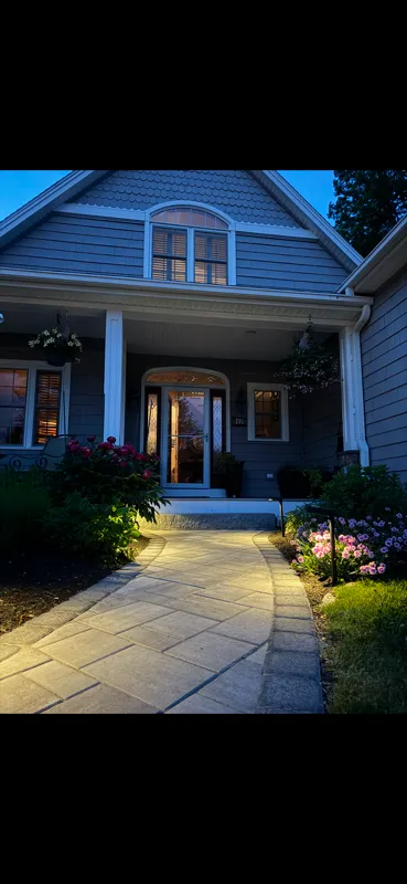 Lit pathway leads to a house with a porch, surrounded by flowers. Blue sky and dark accents.