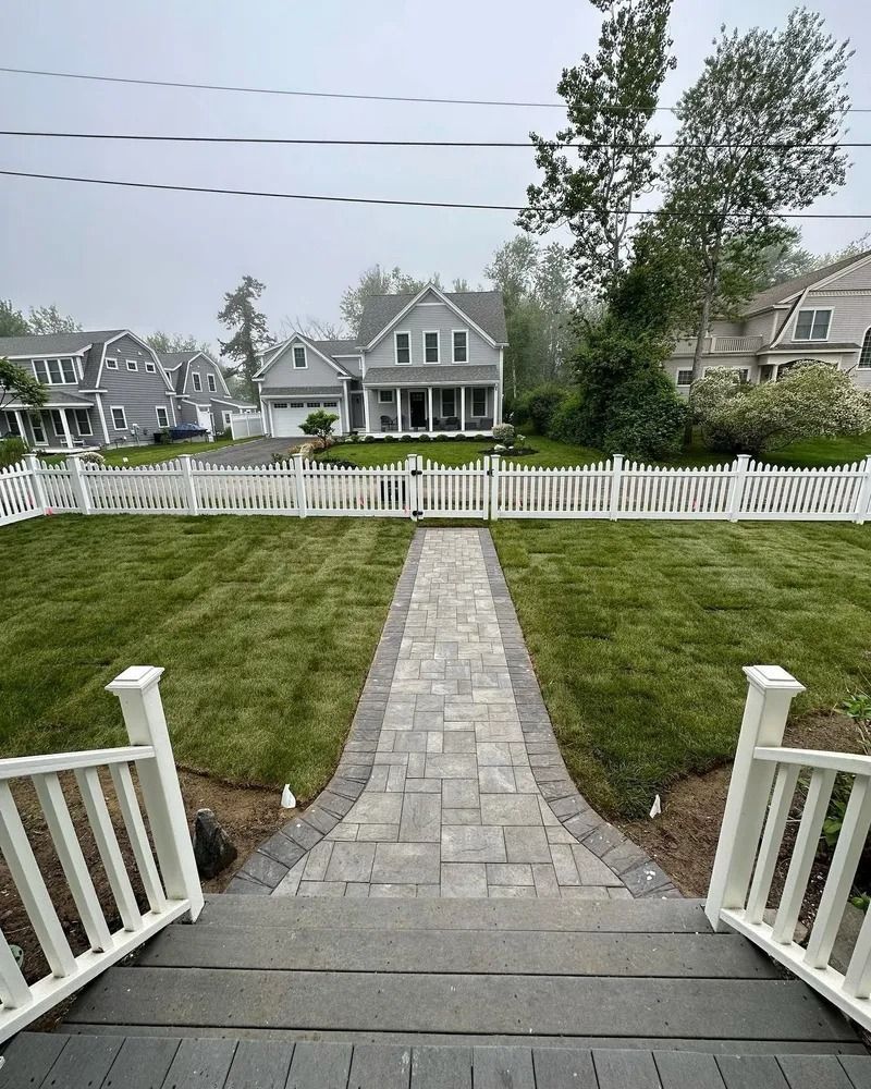 View of a gray house with white picket fence and paved walkway through a green lawn. Foggy.