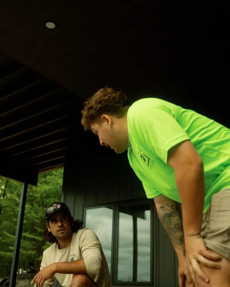 Two men, one in neon green shirt, looking down. The other sits, wearing a tan long sleeve. Exterior setting.