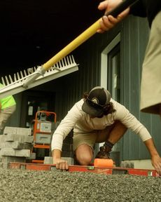 Person laying pavers, kneeling, using a level. Another person holds a rake. Construction setting.