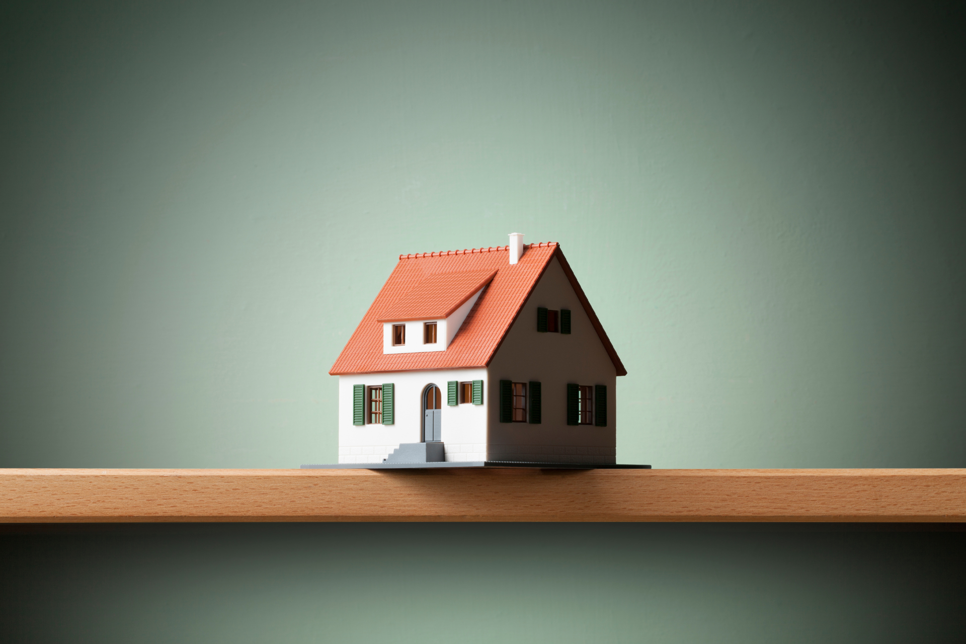 A small model house with a red roof is sitting on a wooden shelf.