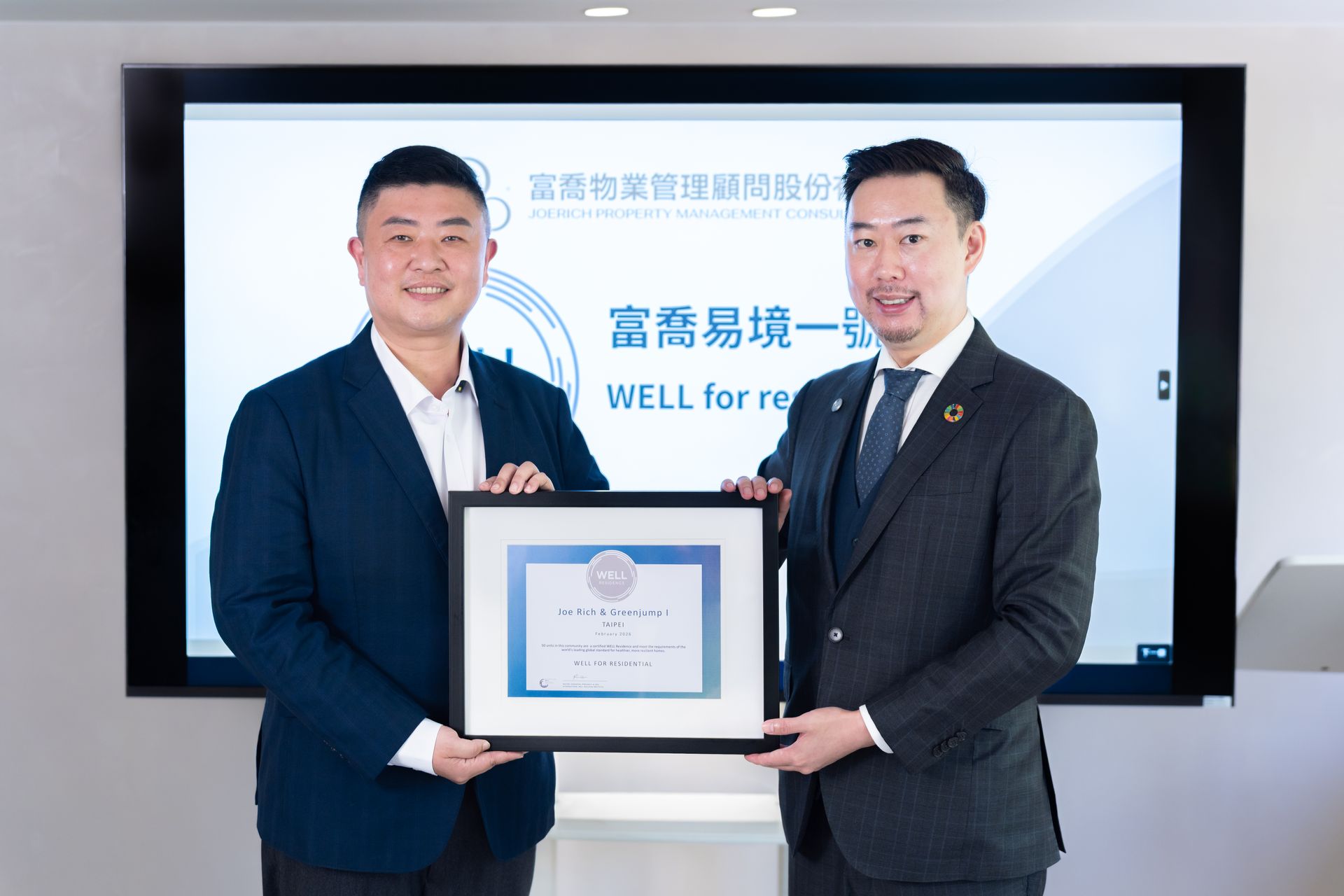 Two men in suits hold a framed certificate in front of a presentation screen.