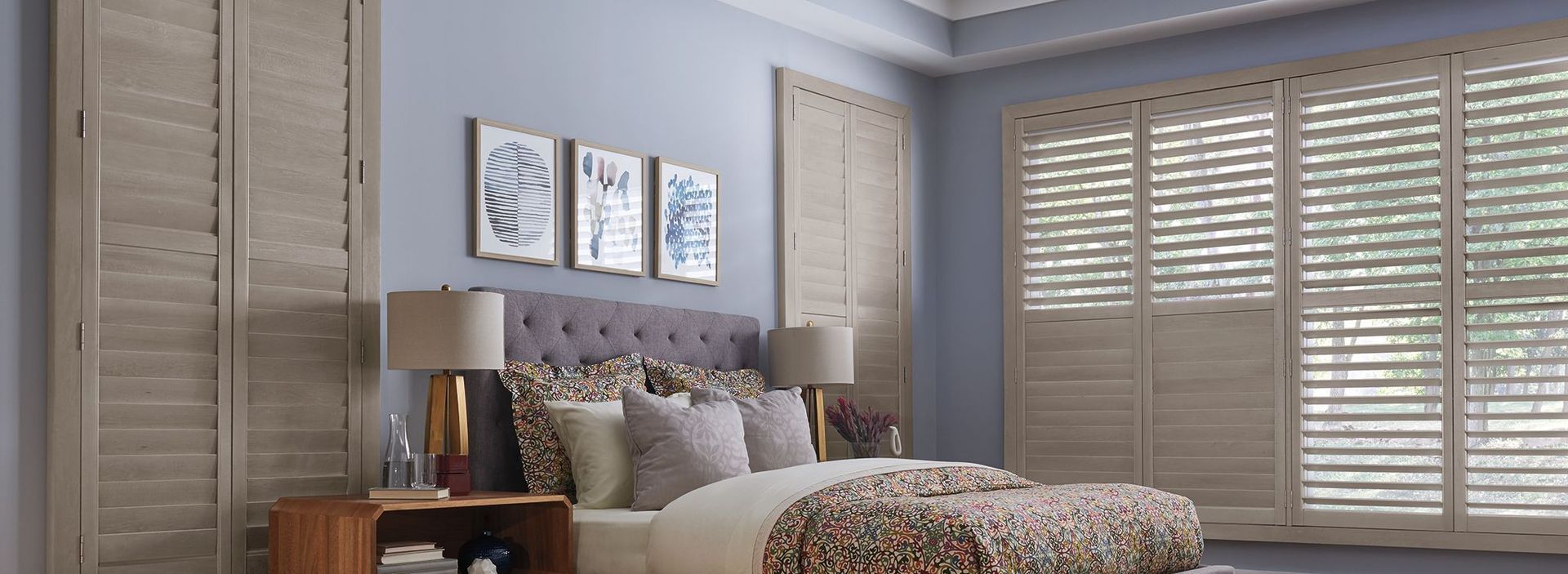 A bedroom with a bed , nightstand , lamps and shutters on the windows.