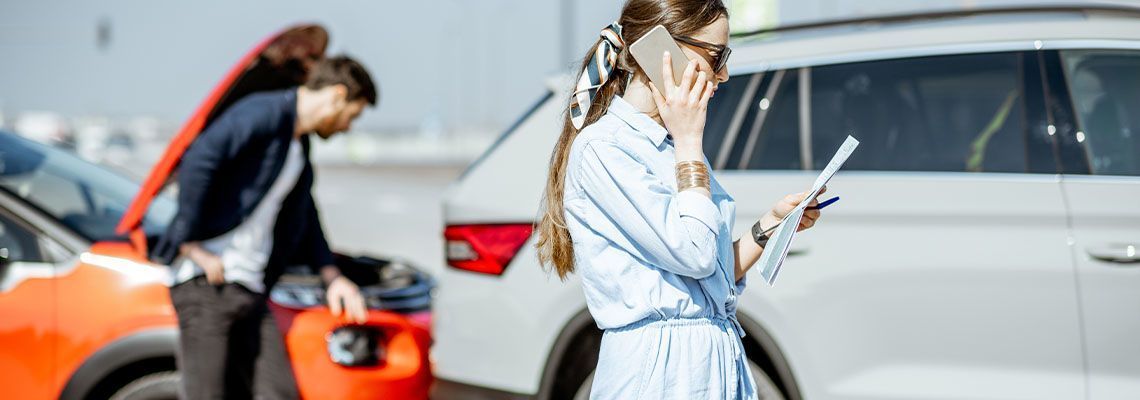 A woman is talking on a cell phone while a man looks under the hood of a car.