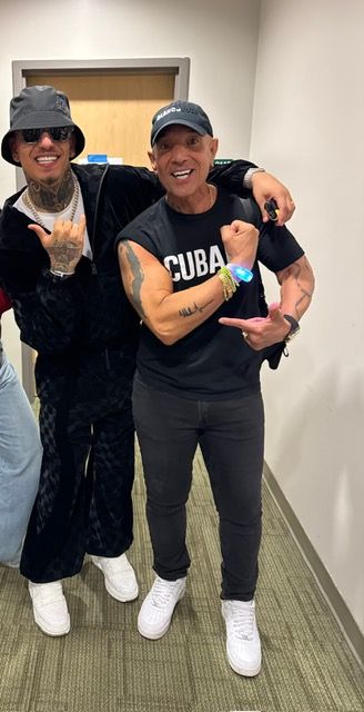 Two men are posing for a picture in a hallway . one of the men is wearing a cuba shirt.