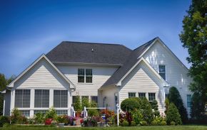 White two-story house with black roof, surrounded by flowers and greenery, under a blue sky.