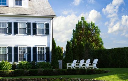White house with black shutters, four white Adirondack chairs on a lawn, blue sky.