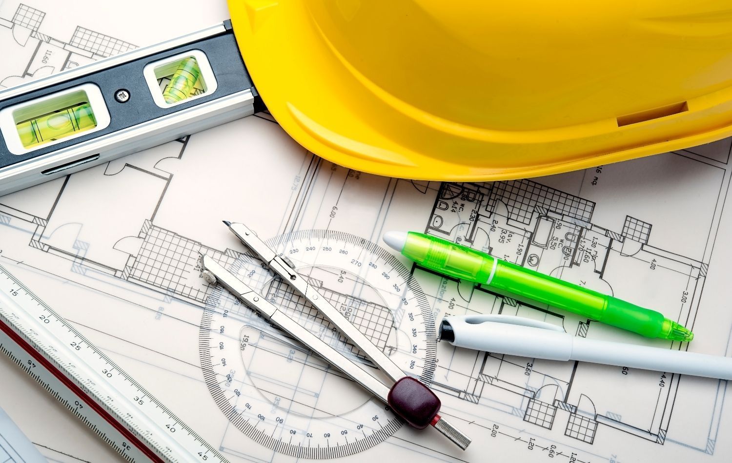 Yellow hard hat, level, drafting tools, and pens on architectural blueprints.