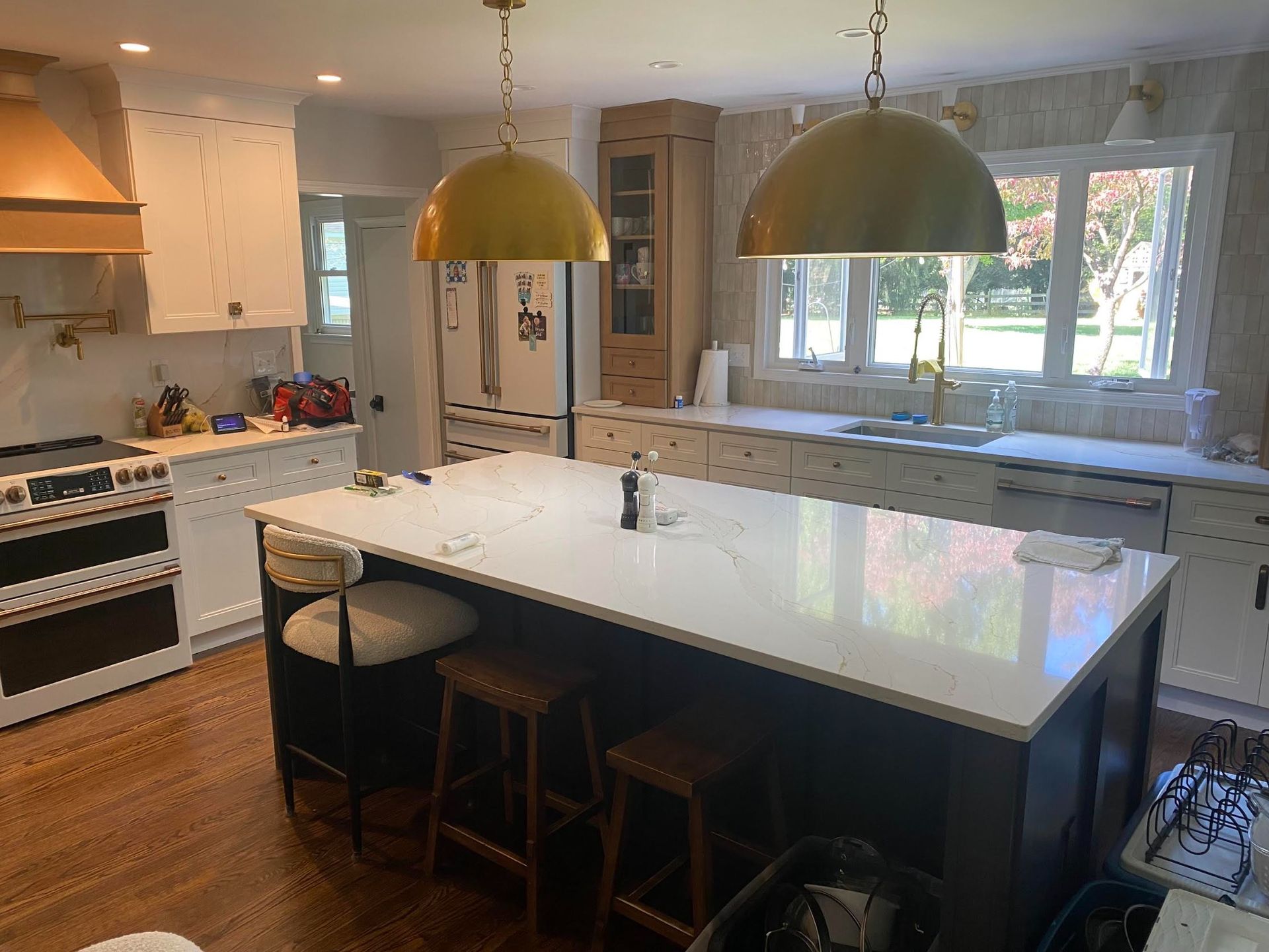 A spacious, bright kitchen with a large dark-based island, white countertops, brass pendant lights, and wooden flooring.