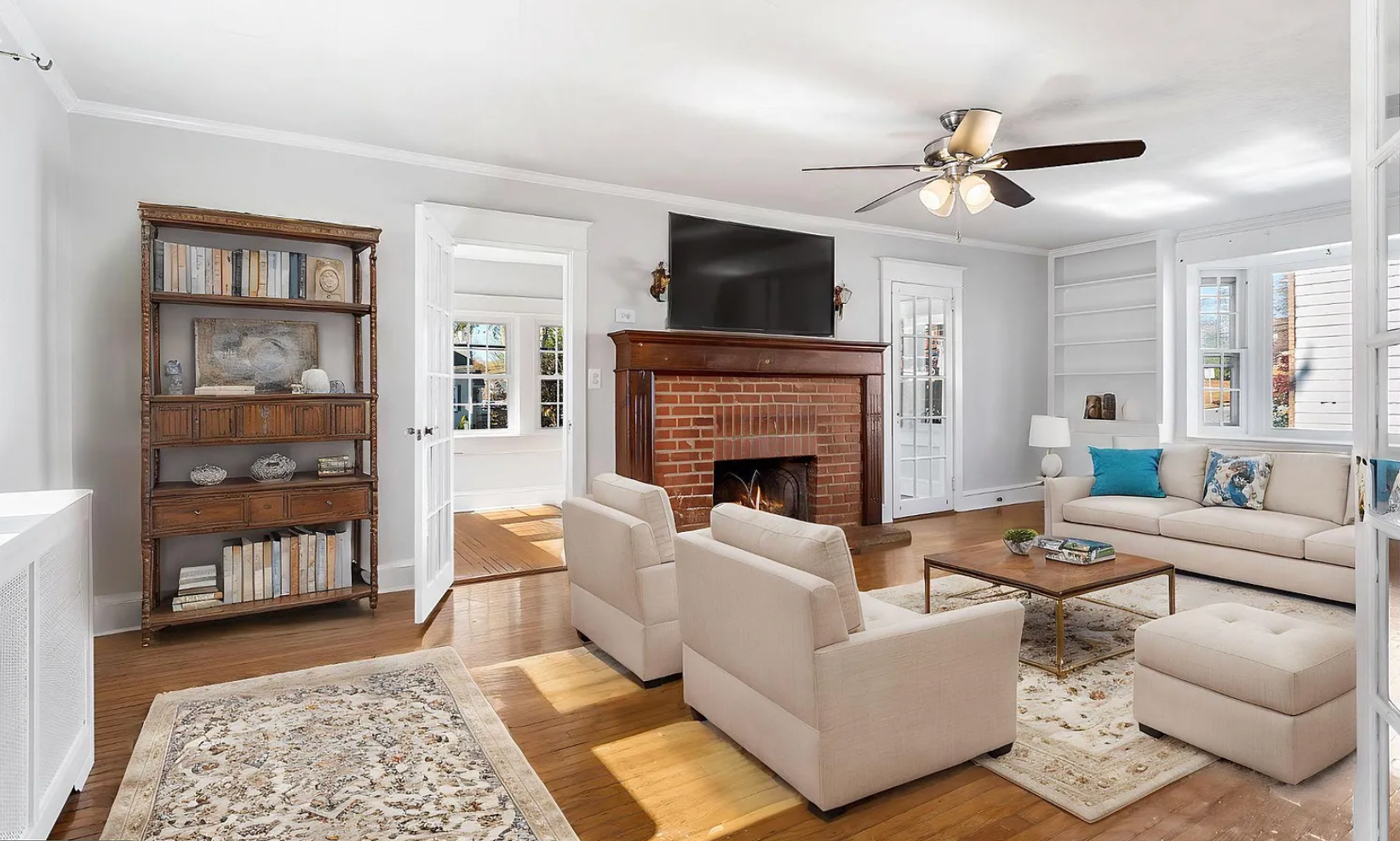 Living room with fireplace, white furniture, bookcase, hardwood floors, and large windows.
